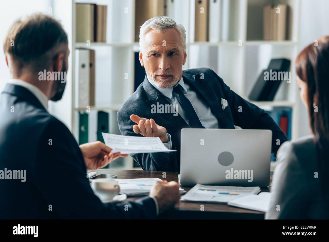 Investisseur mature pointant vers un homme d'affaires avec un document sur un premier plan flou près des papiers et de l'ordinateur portable Banque D'Images