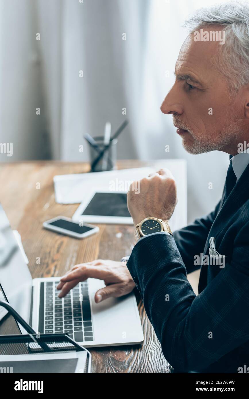 Investisseur en costume utilisant un ordinateur portable près d'appareils sur un arrière-plan flou au bureau Banque D'Images