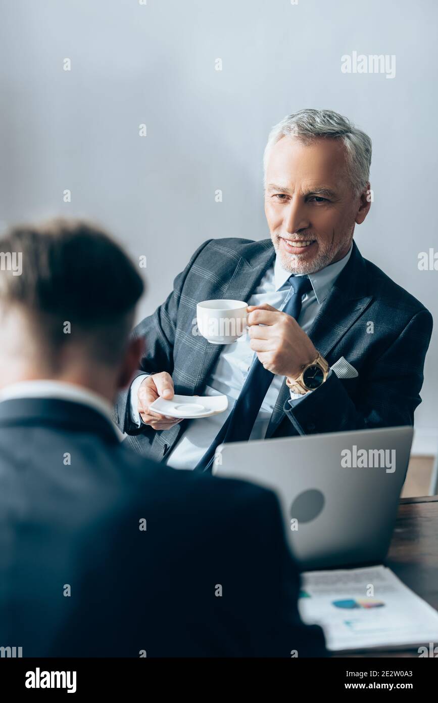 Un investisseur positif tenant une tasse de café près d'un ordinateur portable et d'un homme d'affaires sur un premier plan flou Banque D'Images