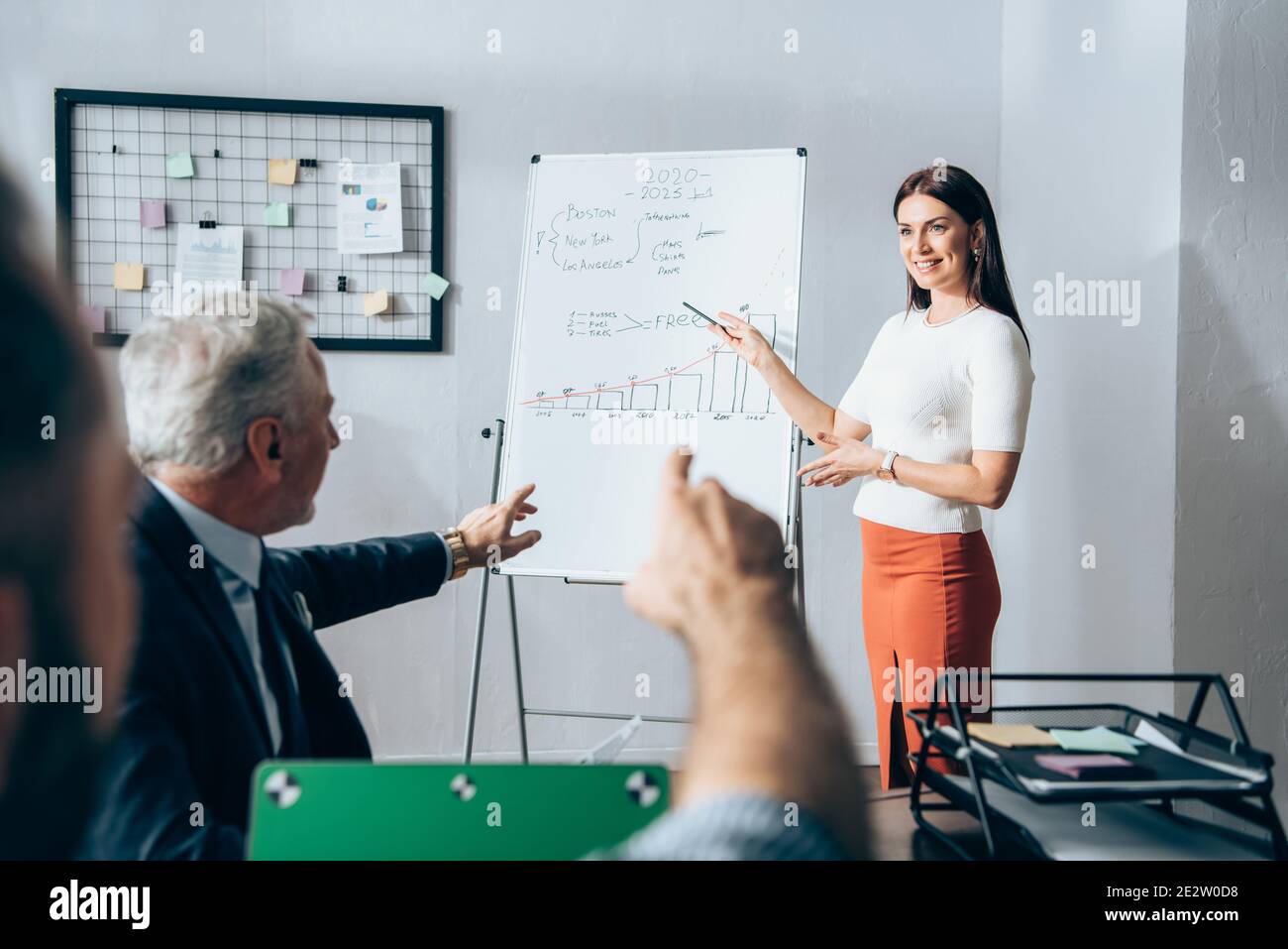 Femme d'affaires souriante pointant sur un tableau de conférence avec des graphiques près de l'investisseur et collègue sur un premier plan flou dans le bureau Banque D'Images