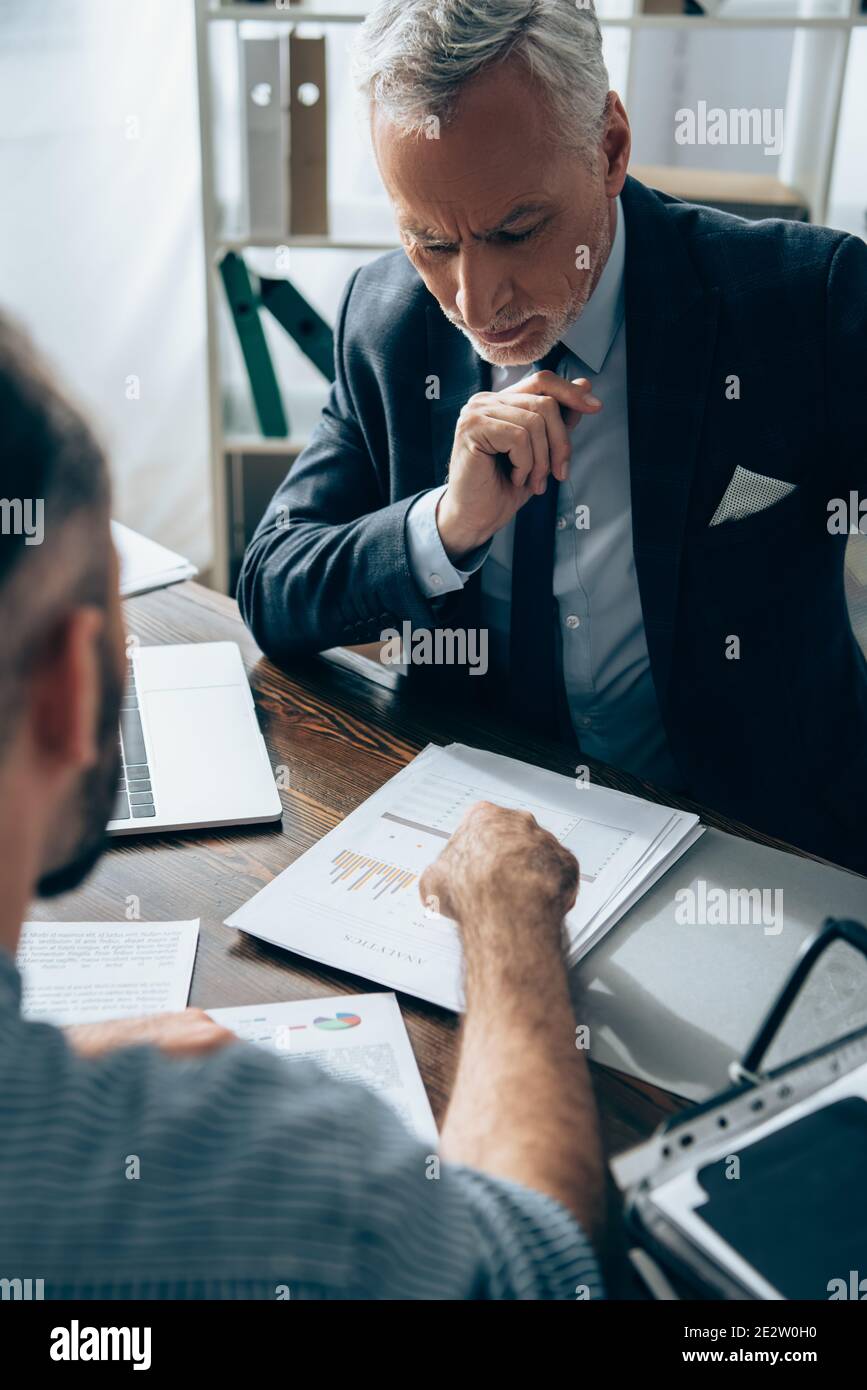 Investisseur pensif regardant des papiers avec des graphiques près d'ordinateur portable et homme d'affaires sur un premier plan flou Banque D'Images