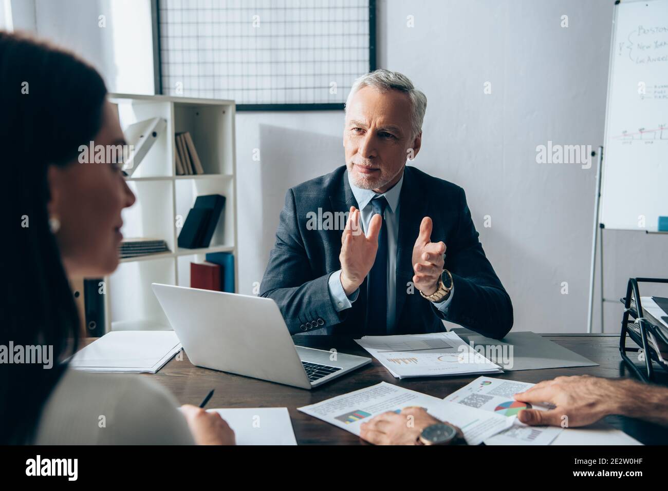 Investisseur en tenue formelle regardant femme d'affaires sur un premier plan flou près de l'ordinateur portable et des documents sur la table Banque D'Images
