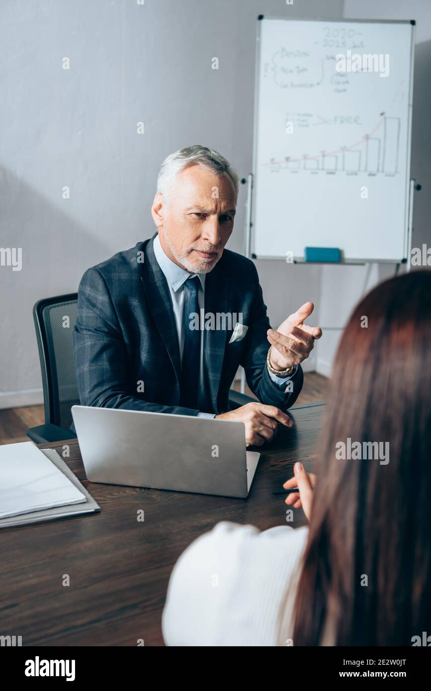 Investisseur mature pointant de la main près d'un ordinateur portable, de papiers et de femme d'affaires sur un premier plan flou Banque D'Images