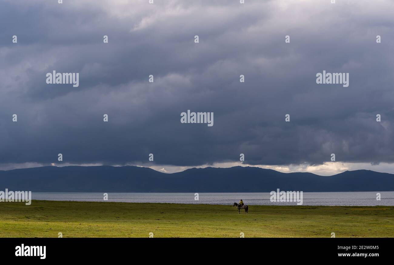 Son Köl, Kirghizistan - 6 juillet 2019 : cavalier solitaire sur un grand pré vert et des nuages brumeux et sombres, Kirghizistan. Banque D'Images Son Köl, Kirghizistan - 6 juillet 2019 : cavalier solitaire sur un grand pré vert et des nuages brumeux et sombres, Kirghizistan. Banque D'Images
