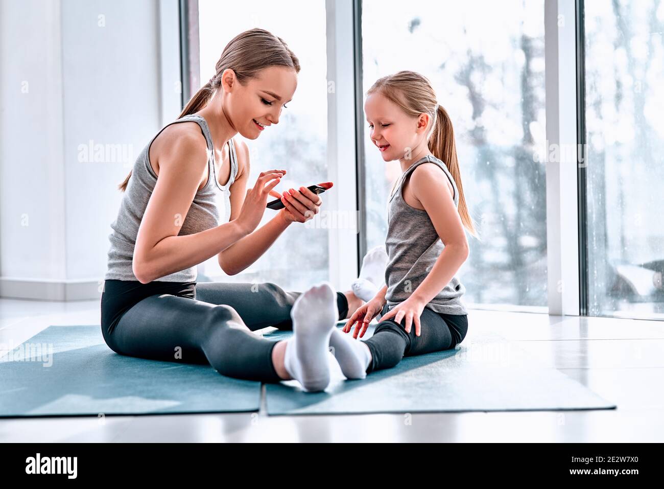 Vue latérale de la jeune femme sportive et de sa petite fille adorable qui a l'air d'un smartphone pour voir comment les jambes s'étirent correctement. Blogueur familial sportif c Banque D'Images