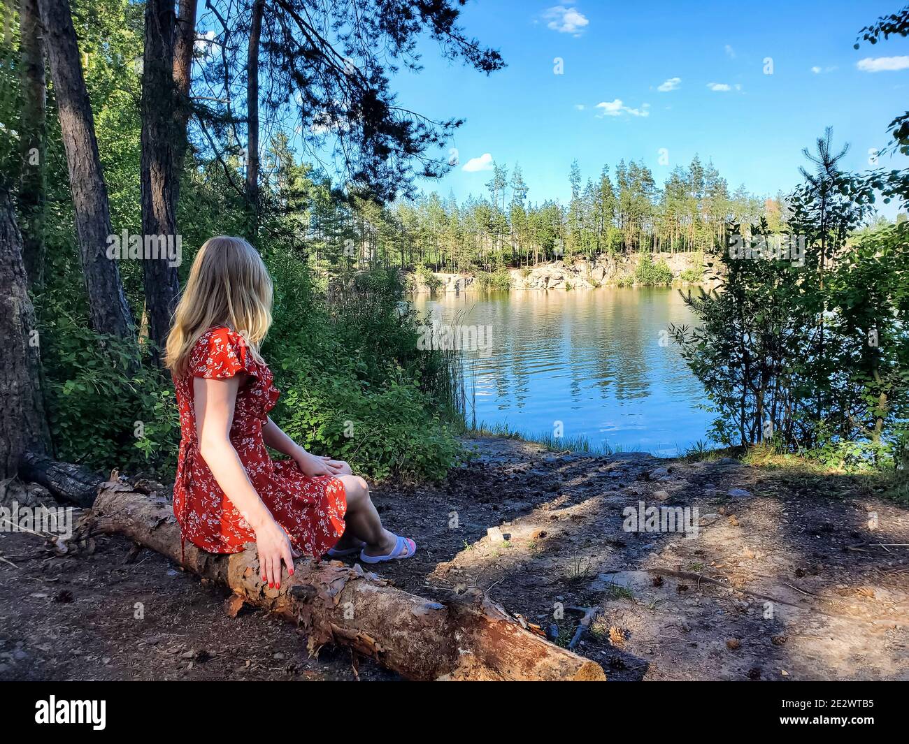 Blonde dans une robe rouge assis sur un arbre tombé sur le rivage d'un lac forestier par temps ensoleillé. Repos campagnard, nature, vue arrière Banque D'Images