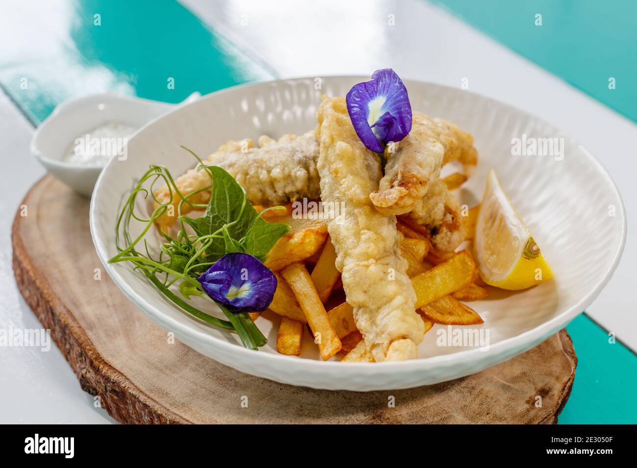 Poisson et frites avec sauce et citron, décoré de fleurs de pois papillons. Servi sur une plaque en céramique blanche. Banque D'Images
