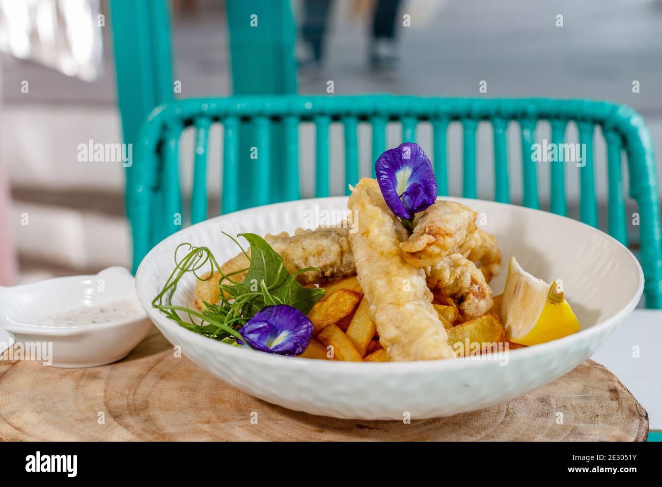 Poisson et frites avec sauce et citron, décoré de fleurs de pois papillons. Servi sur une plaque en céramique blanche. Banque D'Images