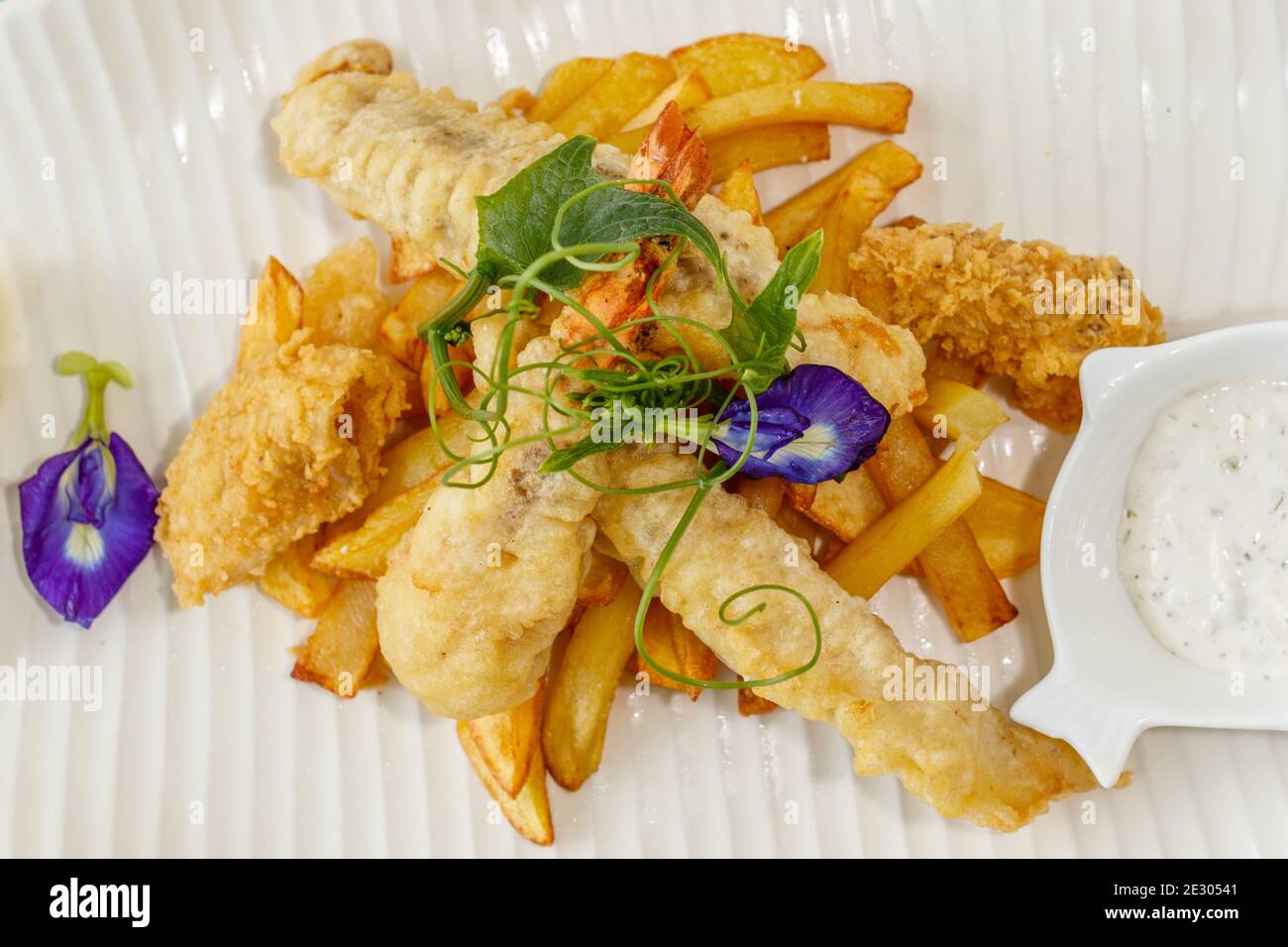 Poisson et frites avec sauce et citron, décoré de fleurs de pois papillons. Servi sur une plaque en céramique blanche. Banque D'Images