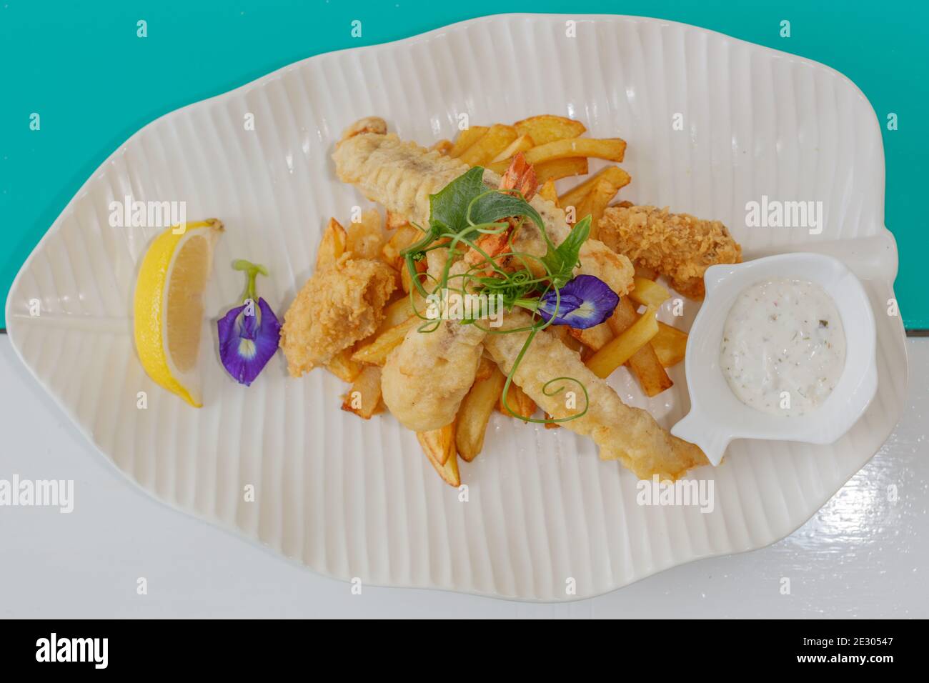 Poisson et frites avec sauce et citron, décoré de fleurs de pois papillons. Servi sur une plaque en céramique blanche. Banque D'Images