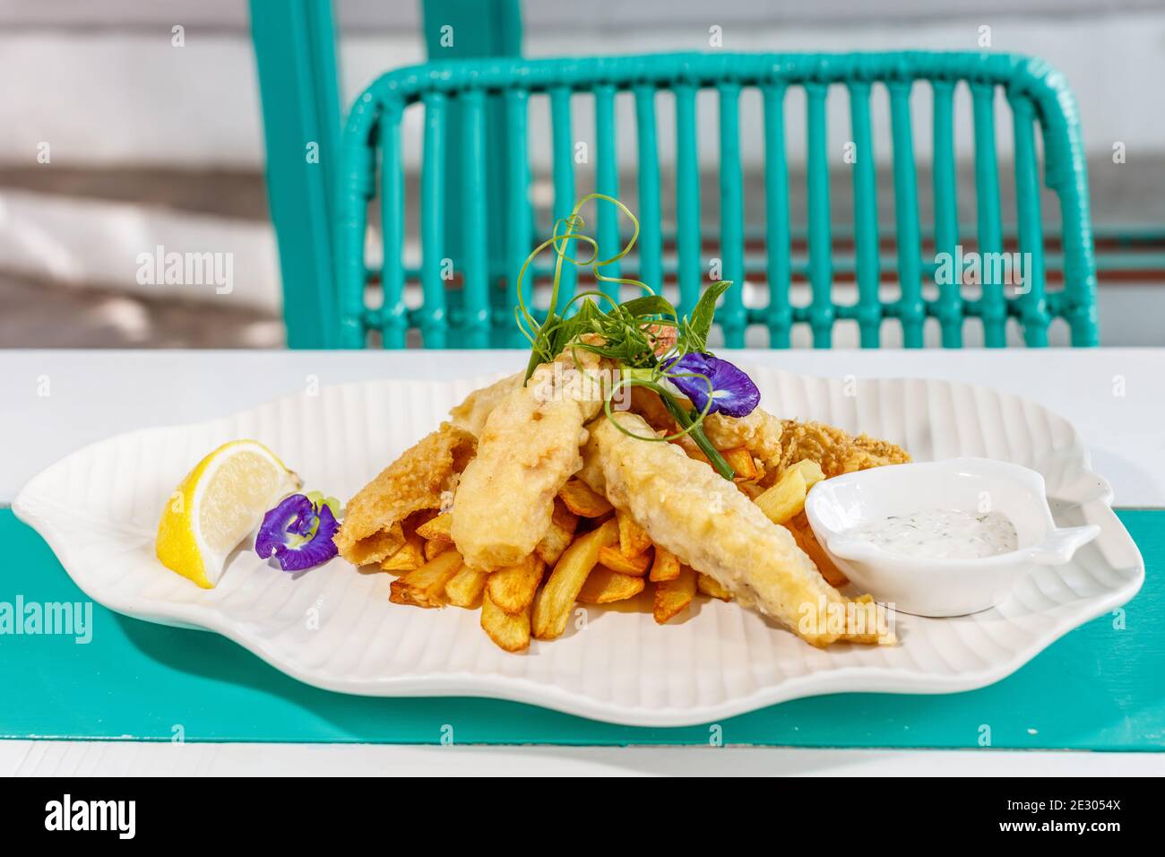 Poisson et frites avec sauce et citron, décoré de fleurs de pois papillons. Servi sur une plaque en céramique blanche. Banque D'Images