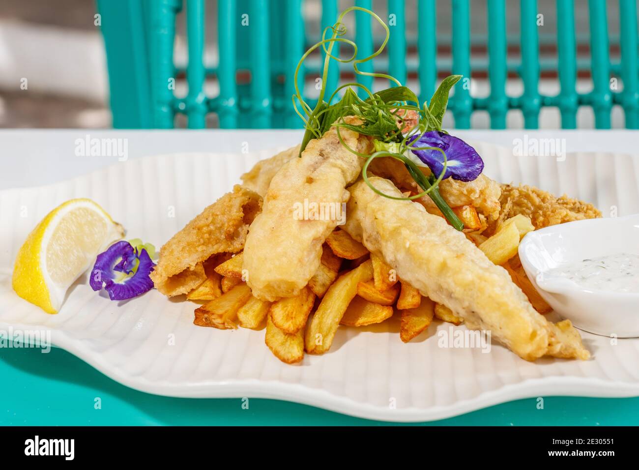 Poisson et frites avec sauce et citron, décoré de fleurs de pois papillons. Servi sur une plaque en céramique blanche. Banque D'Images