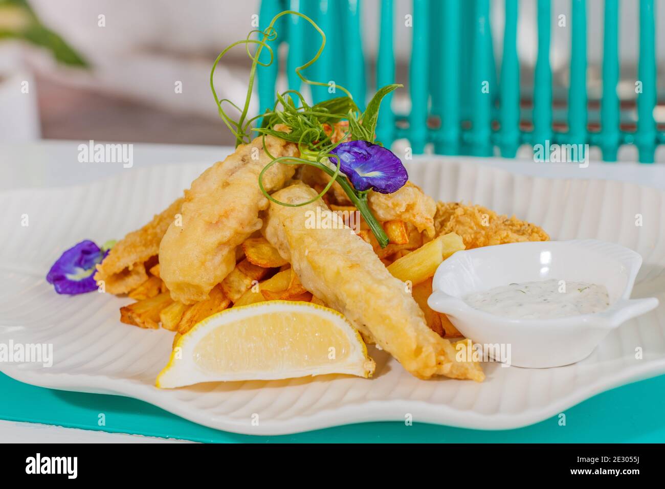 Poisson et frites avec sauce et citron, décoré de fleurs de pois papillons. Servi sur une plaque en céramique blanche. Banque D'Images