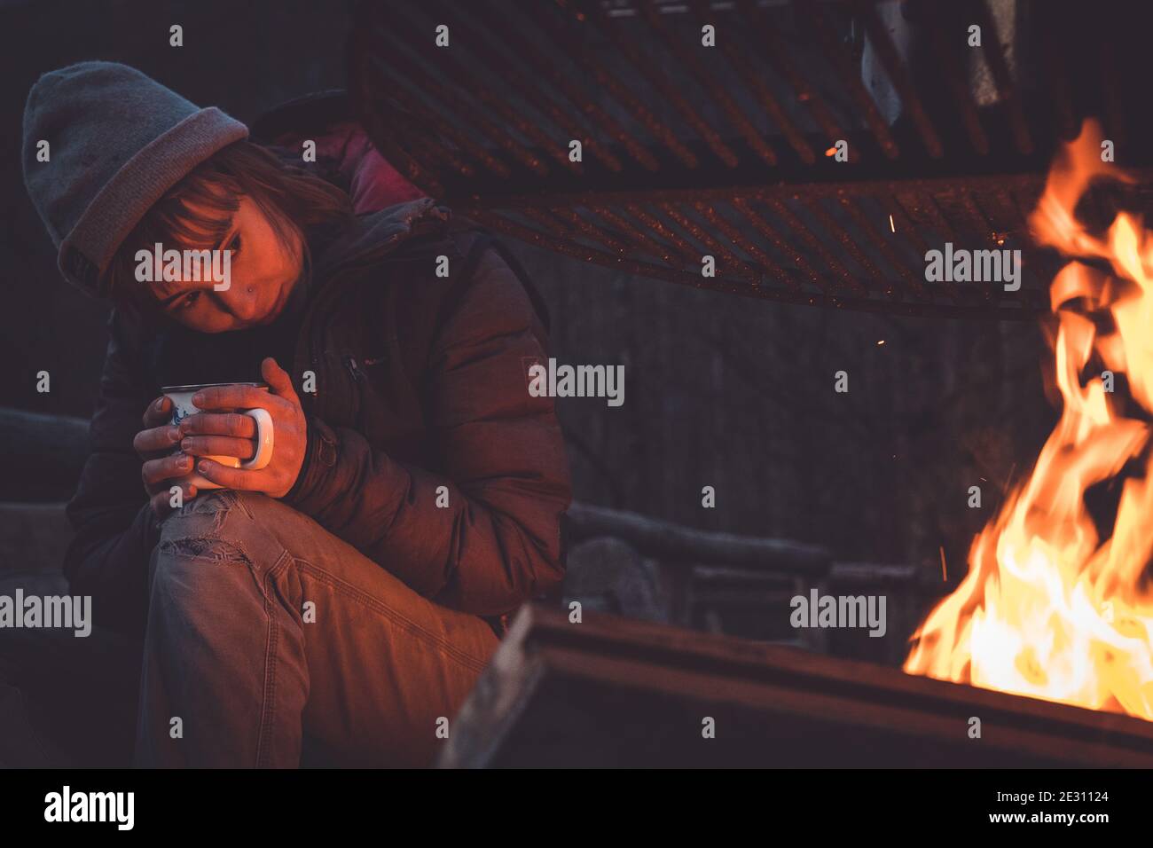 Une femme qui tient une boisson chaude regarde dans les flammes d'un feu de camping Banque D'Images