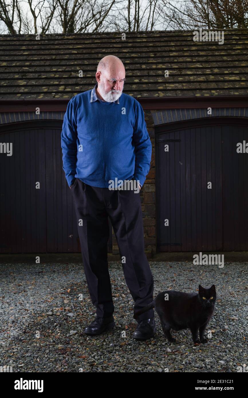 Homme âgé debout devant son garage avec son chat noir errant passé. Banque D'Images