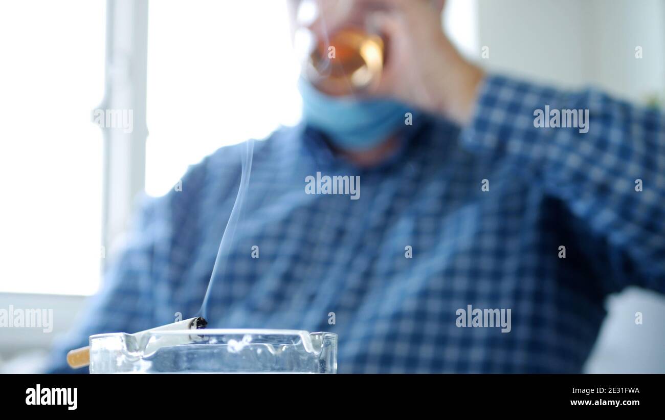 Image avec un homme qui boit de l'alcool et fume un Cigarette portant un masque de protection sur son visage Banque D'Images