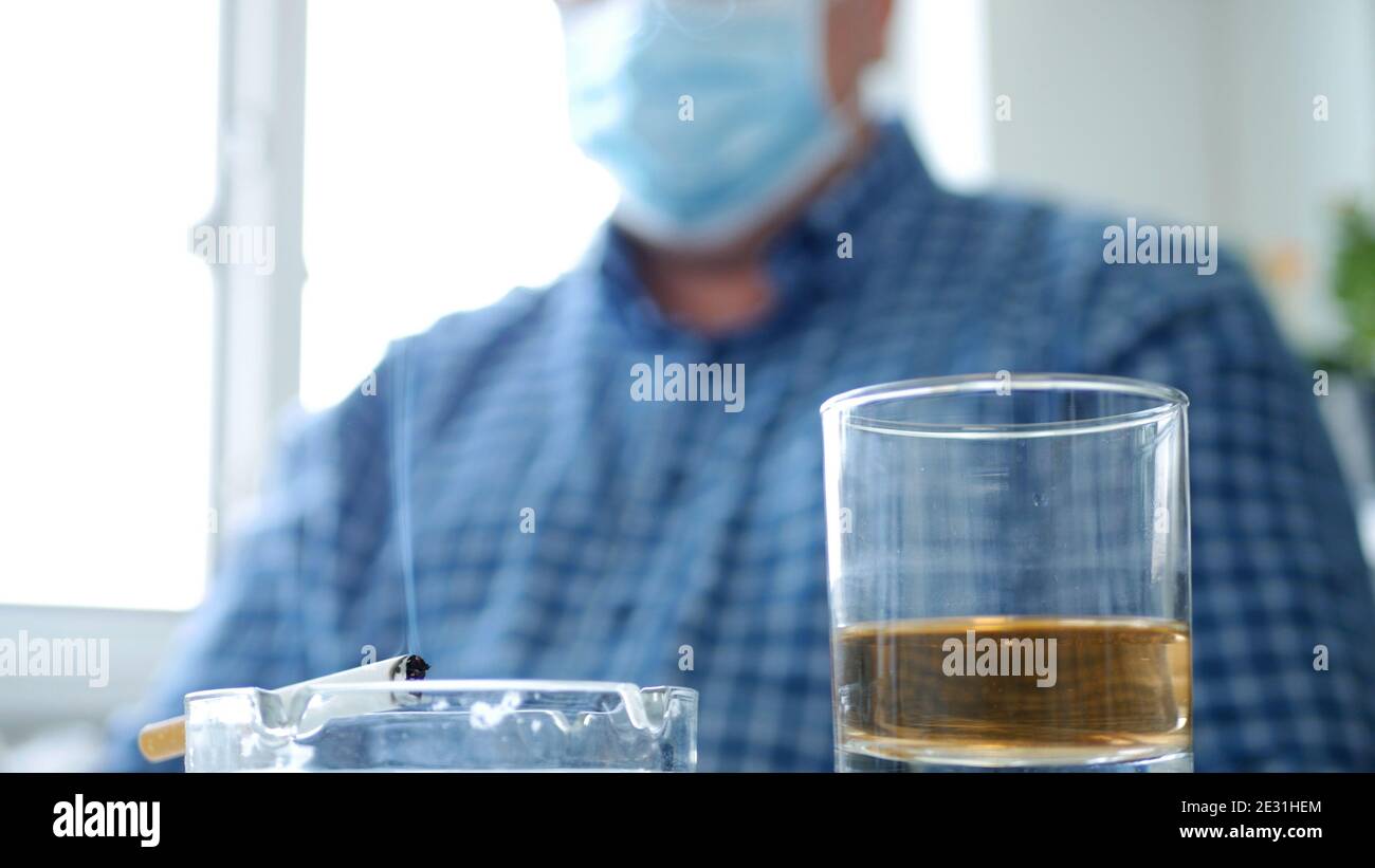 Image avec un homme qui boit de l'alcool et fume un Cigarette portant un masque de protection sur son visage Banque D'Images