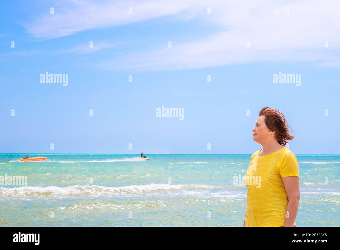 Une femme âgée dans un T-shirt jaune se tient sur le rivage de la mer d'azur contre un ciel bleu, regarde dans la distance et apprécie une journée d'été, l'ope Banque D'Images