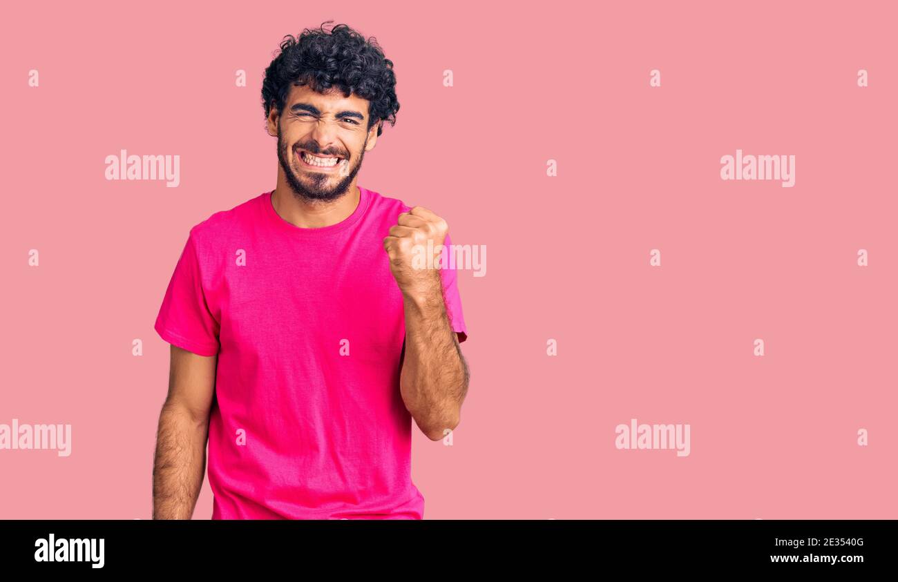 Beau jeune homme aux cheveux bouclés et ours portant un tshirt rose décontracté en colère et fou levant le poing frustré et furieux tout en criant avec colère. ra Banque D'Images