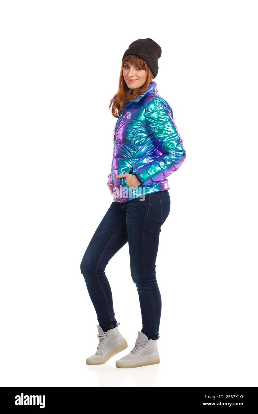 Belle jeune femme en duvet, casquette d'hiver, jeans et baskets, les mains dans la poche et souriant. Long studi Banque D'Images