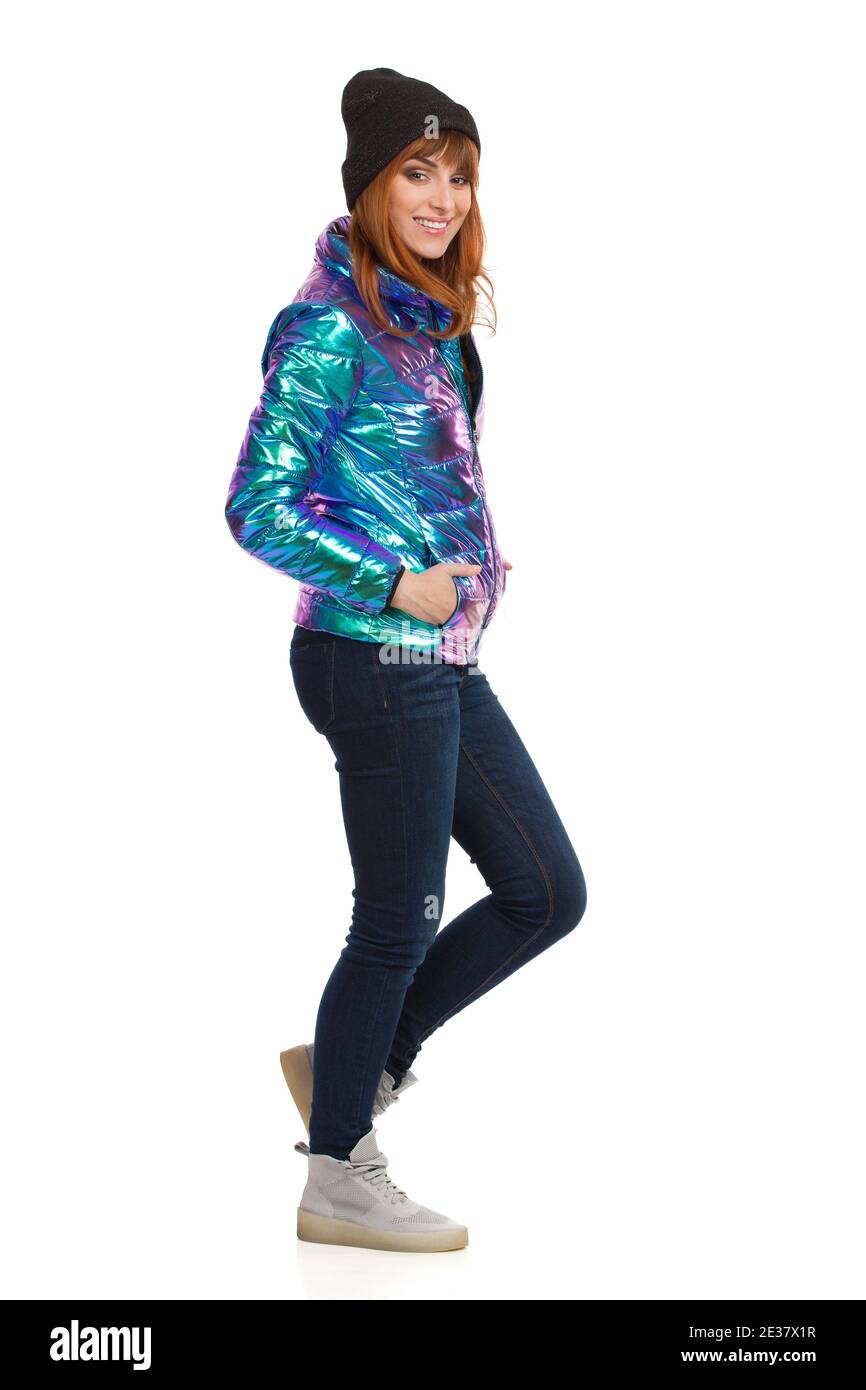 Jeune femme en duvet, jeans, baskets et casquette brillants et vibrants, debout, tenant la main dans la poche et souriant. Vue latérale. Studios pleine longueur s Banque D'Images