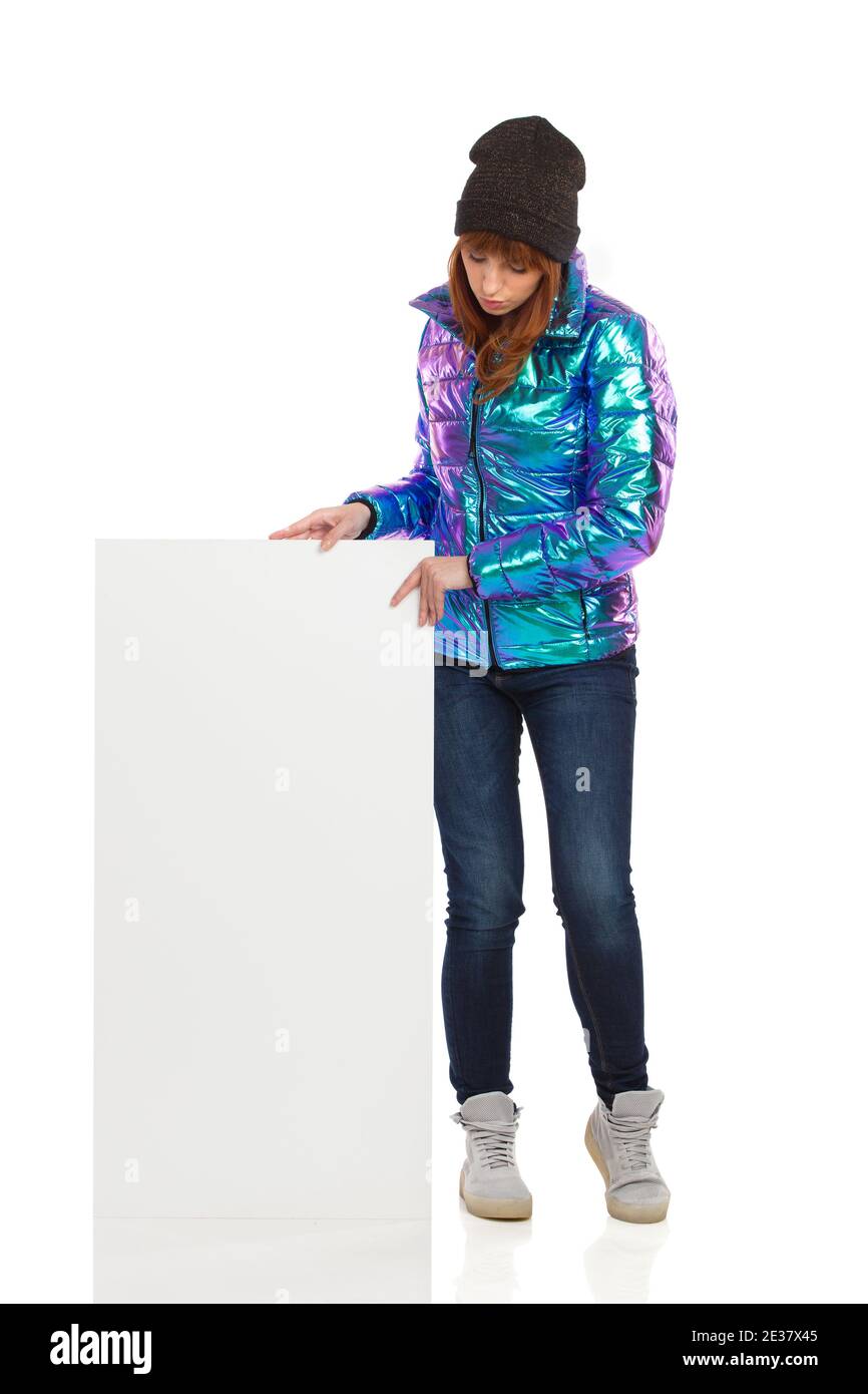 Jeune femme en duvet, casquette d'hiver, jeans et baskets est debout, avec un écriteau blanc, regardant vers le bas et lisant. Longueur totale Banque D'Images