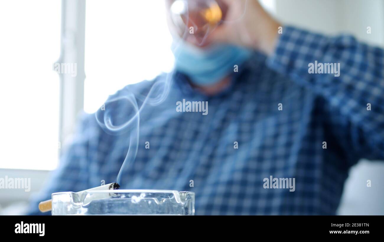 Image avec un homme qui boit de l'alcool et fume un Cigarette portant un masque de protection sur son visage Banque D'Images