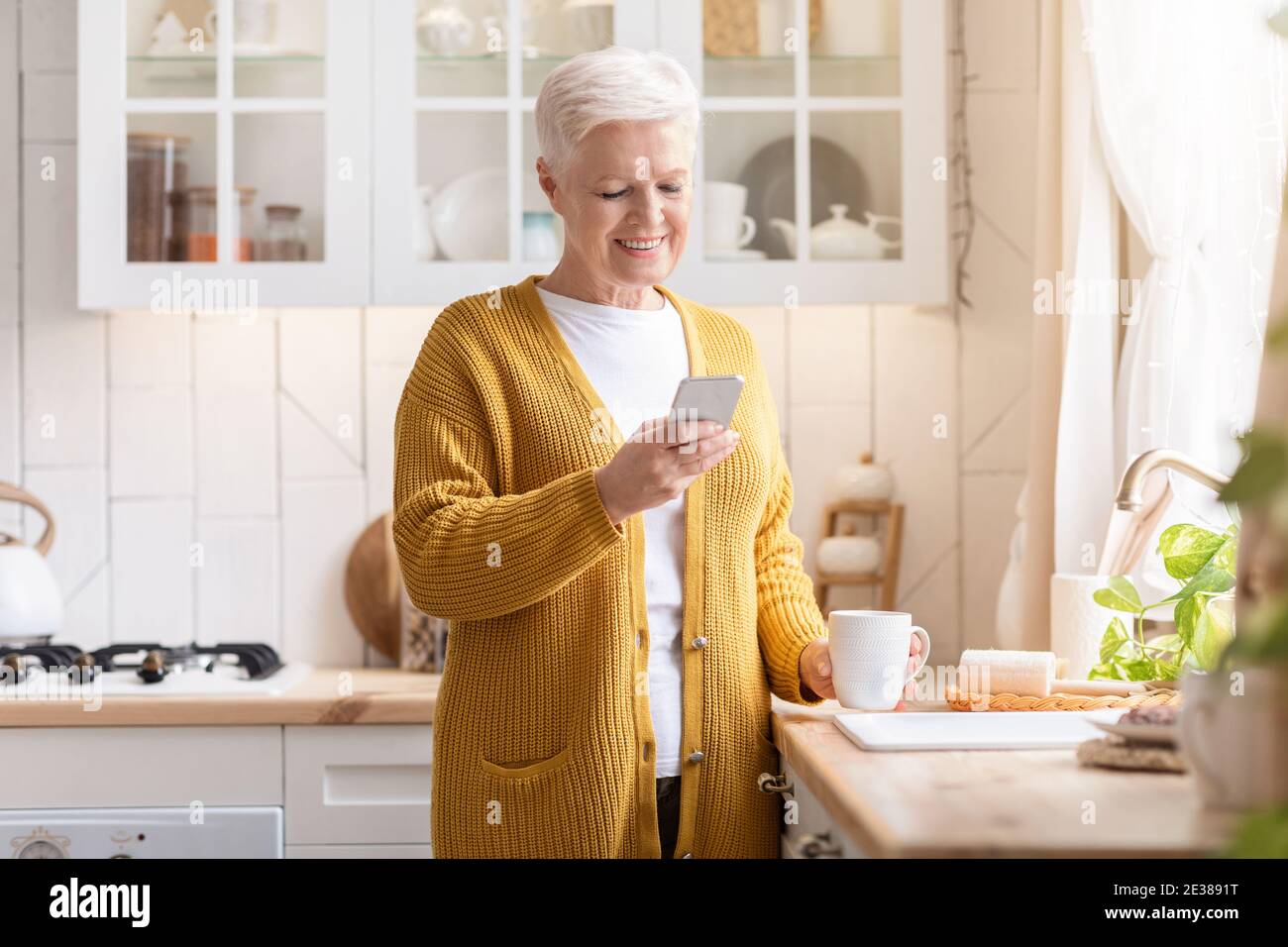 Une vieille dame souriante utilisant un téléphone portable tout en buvant du café Banque D'Images