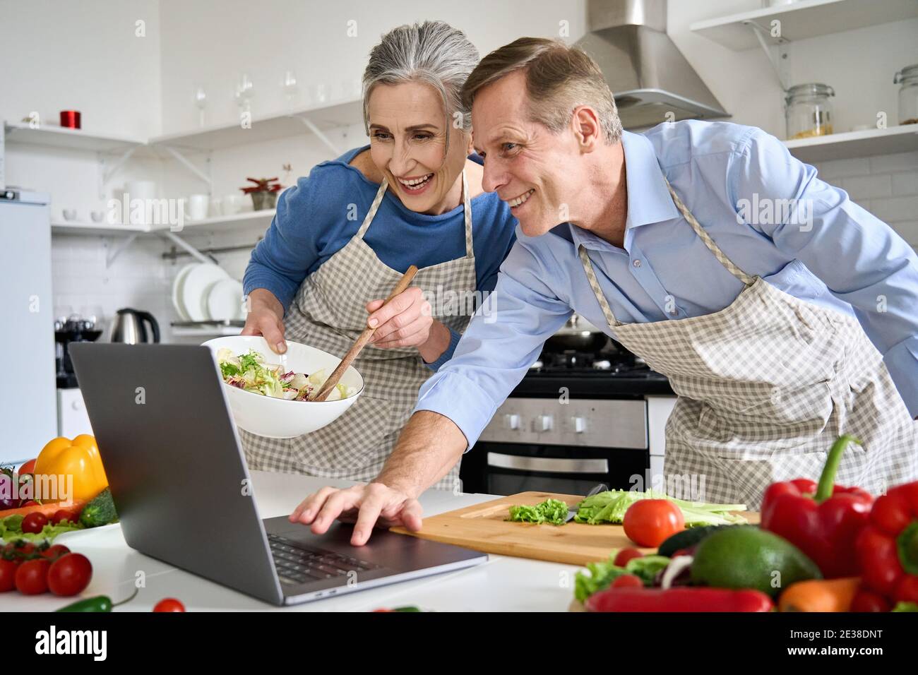 Un vieux couple heureux utilisant un ordinateur portable pour préparer une salade végétalienne à la maison dans la cuisine. Banque D'Images