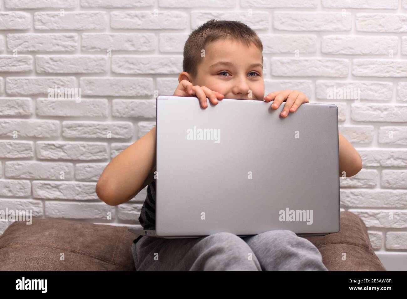 Un garçon de 6 à 7 ans est assis sur le sol avec un ordinateur portable. Contre un mur de briques. L'enfant regarde derrière l'ordinateur portable. Banque D'Images