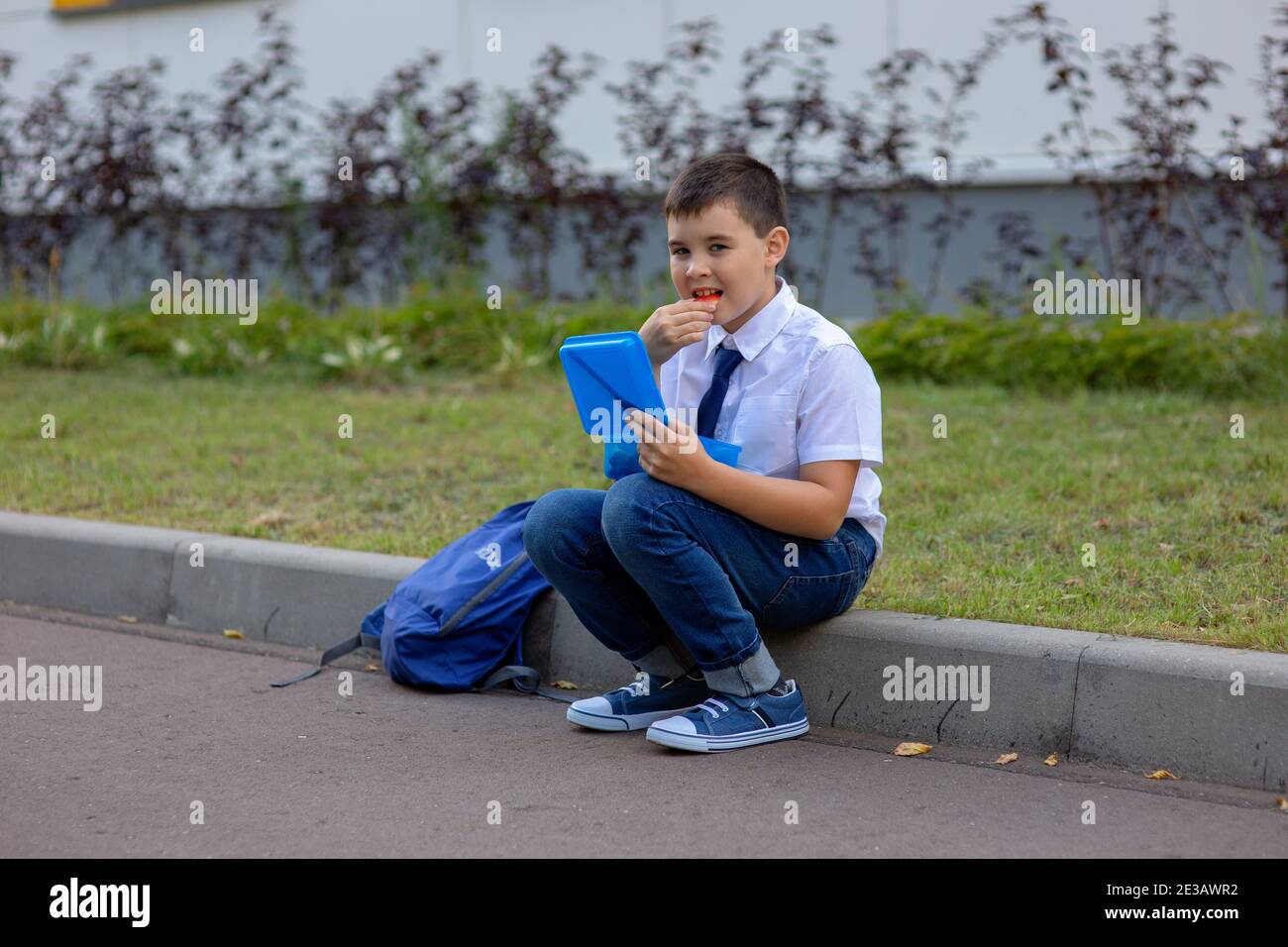 un écolier dans une chemise blanche avec une cravate bleue tient une boîte à lunch bleue et mange un morceau de pomme Banque D'Images