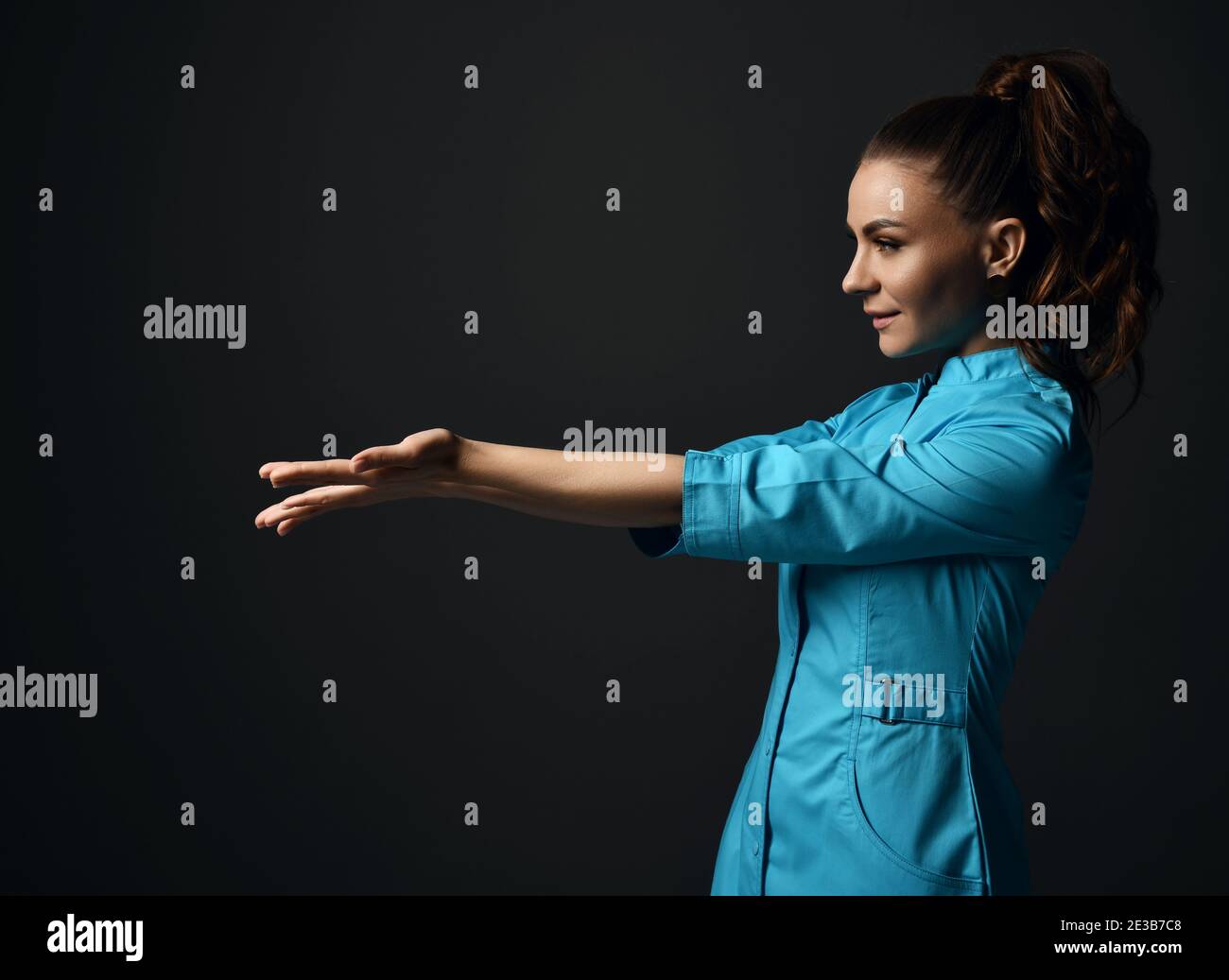 Jeune femme brune médecin infirmière en robe médicale bleue tient ses mains avec des paumes ouvertes avec l'espace de copie. Vue latérale Banque D'Images