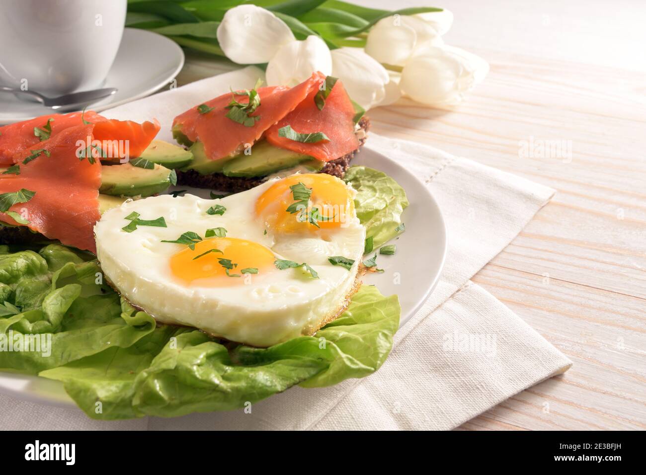 Petit déjeuner de vacances avec amour, œufs frits en forme de coeur, sandwichs avec avocat et saumon fumé sur laitue, tulipes blanches sur un t de bois peint clair Banque D'Images