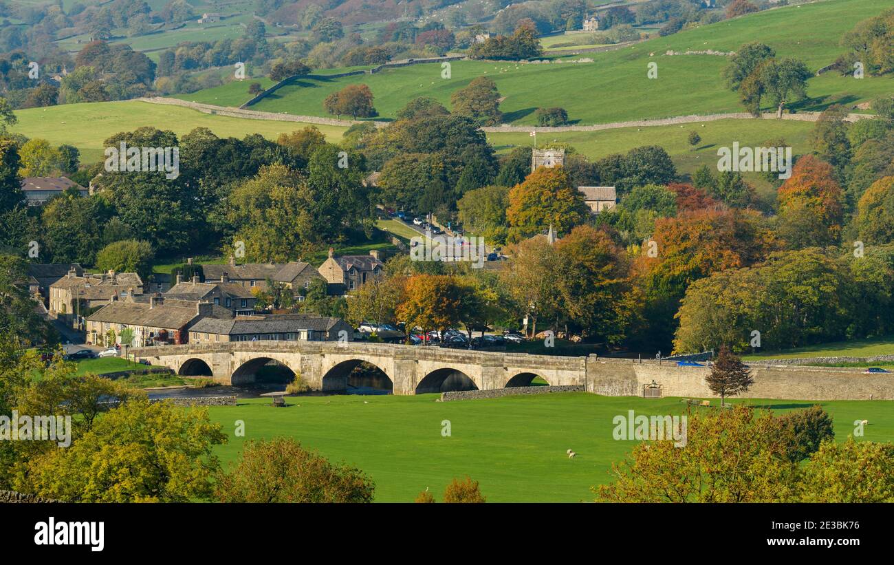 Pittoresque village ensoleillé de Burnsall (pont en pierre de 5 arcades, rivière Wharfe, chalets, église, champs de flanc de colline, arbres d'automne) - Yorkshire Dales, Angleterre Royaume-Uni. Banque D'Images