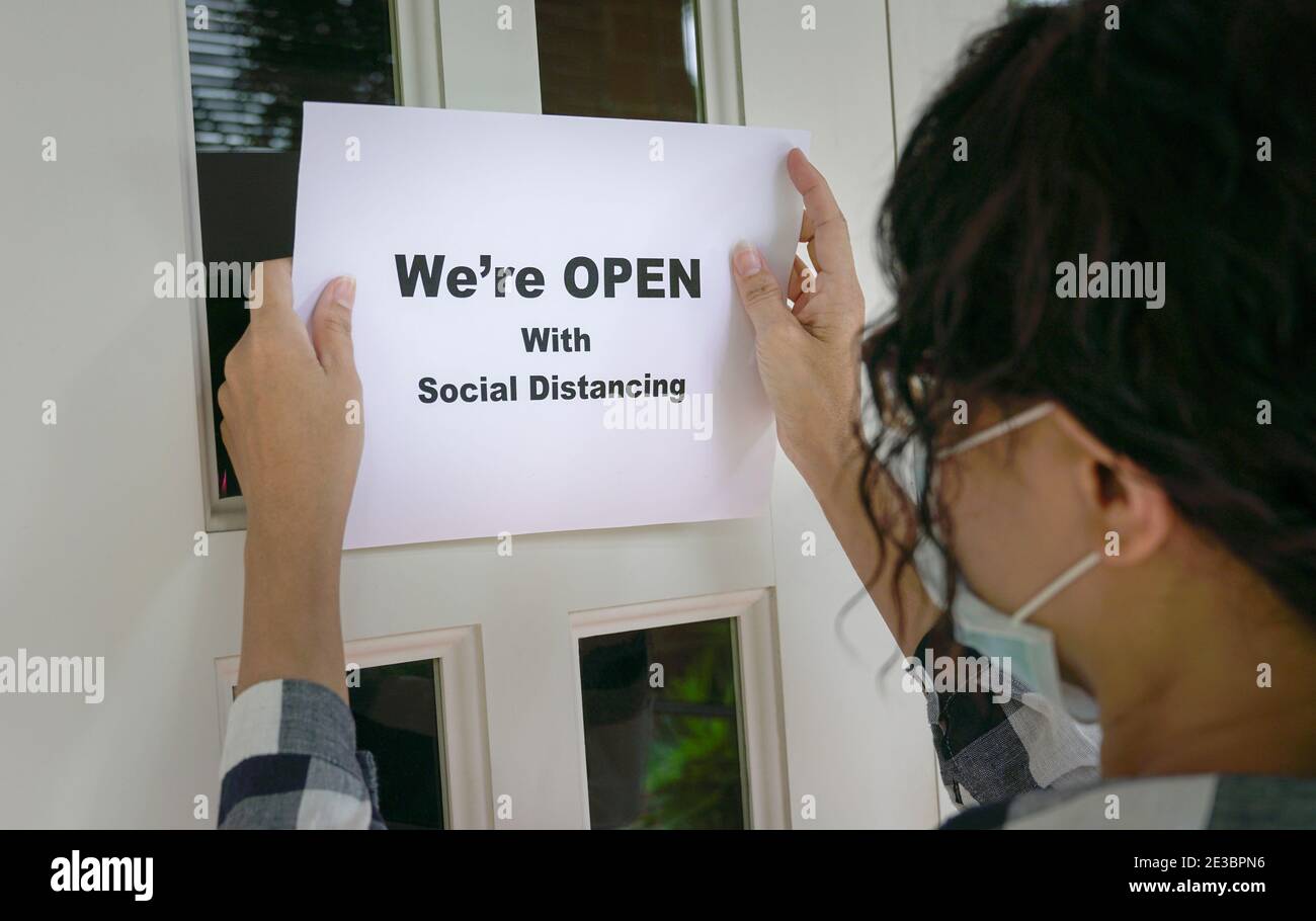 Femme portant un masque de visage mettant un avis, nous sommes ouverts avec social distancer sur la porte d'entrée. Les opérations commerciales avec la nouvelle norme au cours de Covid-19 pand Banque D'Images