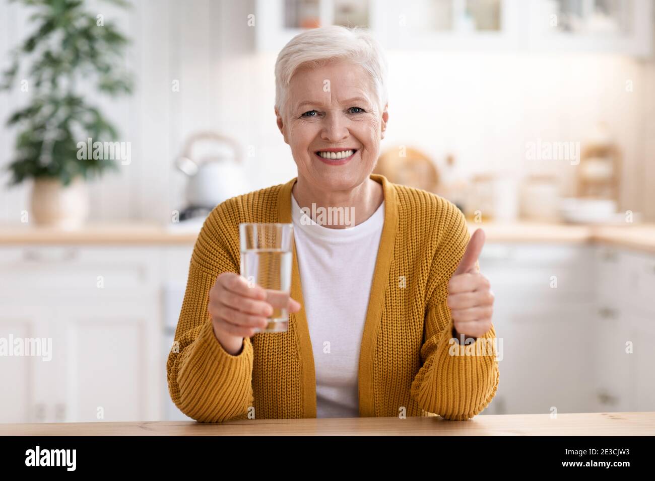 Femme aîée gaie avec un verre d'eau montrant le pouce vers le haut Banque D'Images