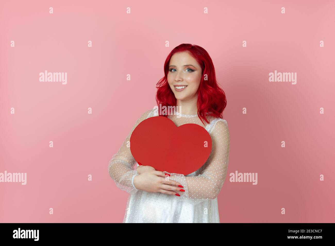 une femme heureuse et souriante en robe blanche et avec des cheveux rouges tenant un grand coeur rouge de papier isolé sur fond rose Banque D'Images