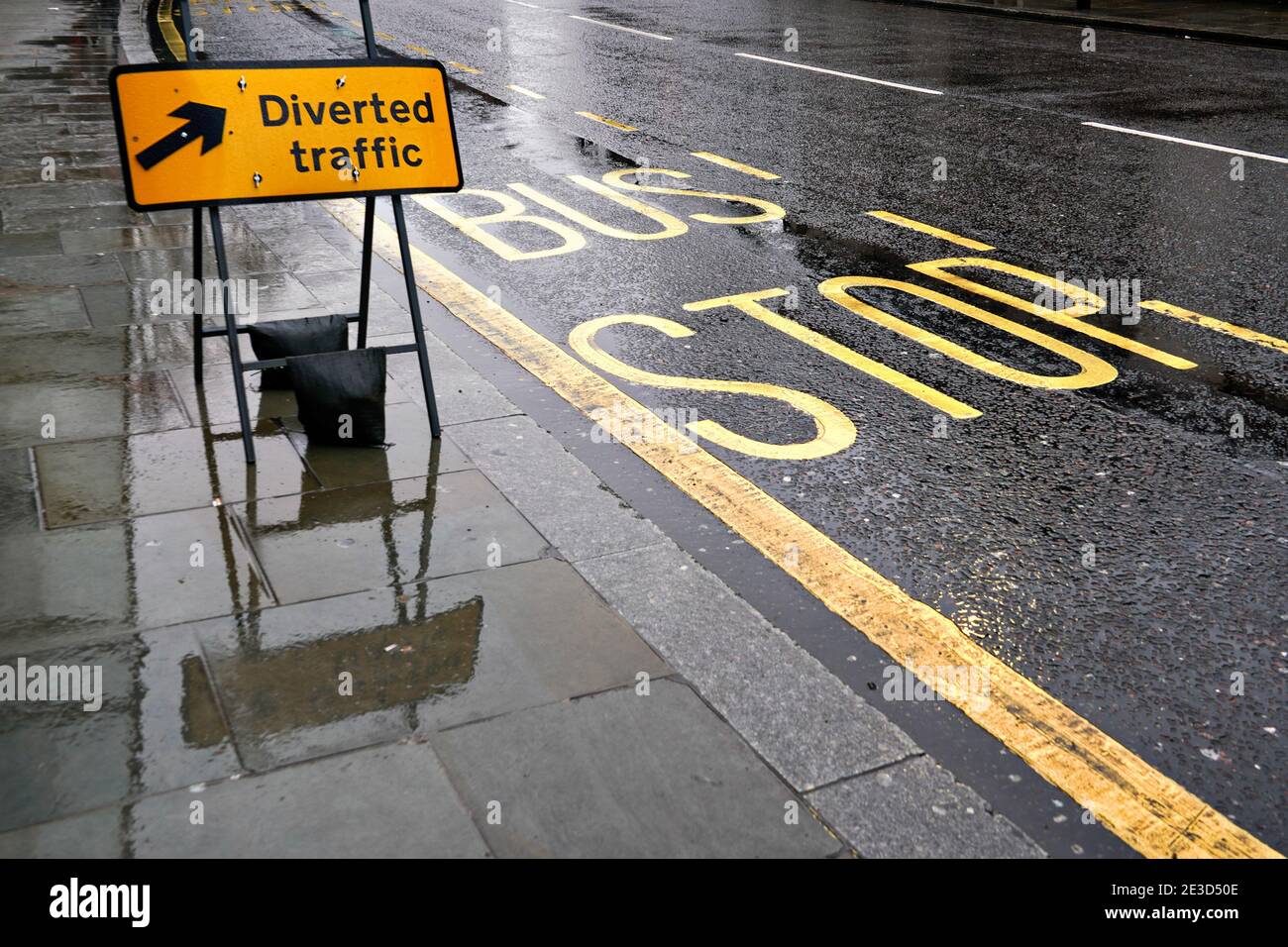 Signalisation noire et jaune sur chaussée mouillée, à côté de la route avec texte d'arrêt de bus, les feux se reflètent dans l'asphalte recouvert de pluie Banque D'Images