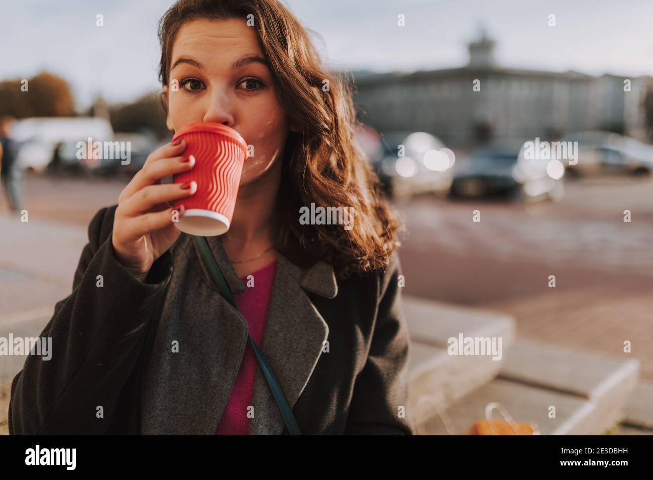 Gros plan de la jolie jeune femme debout dans la ville tout en dégustant un café avec des voitures en arrière-plan Banque D'Images