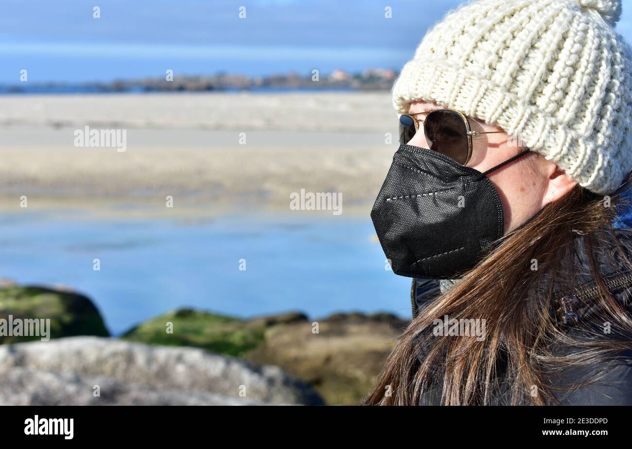 Femme avec masque noir Covid-19 sur une plage en hiver avec manteau noir,  lunettes de soleil et chapeau de laine blanche. Masque KN95 ou N95 ou FFP2  Photo Stock - Alamy