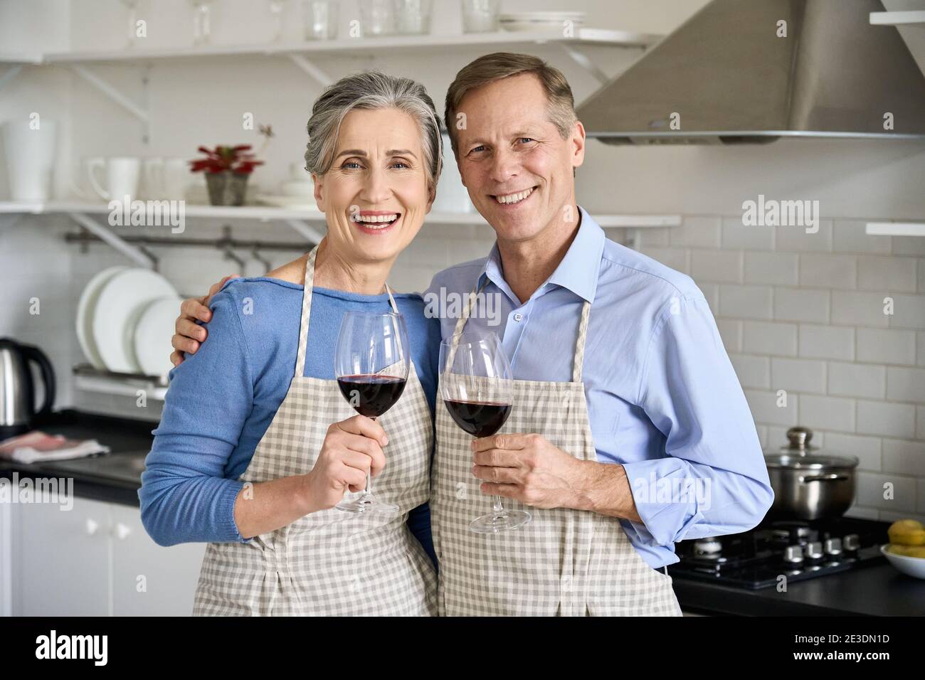 Joyeux vieux couple des années 50 qui s'embrasse en buvant du vin, regardant l'appareil photo dans la cuisine. Banque D'Images