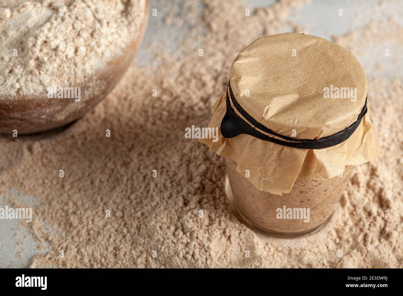 Ingrédients faits maison pour faire du pain au levain avec un bol de farine et une culture de base de levain dans une tasse de verre couverte avec un papier marron sur le dessus Banque D'Images