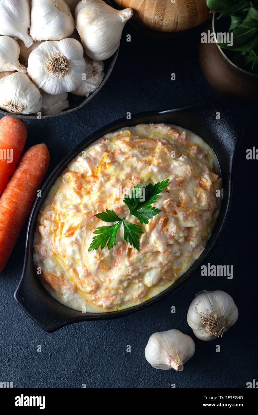 Tarateur de carottes dans un bol en céramique avec légumes sur fond sombre. Salade de carottes fraîche avec yaourt à l'ail dans un bol. Banque D'Images