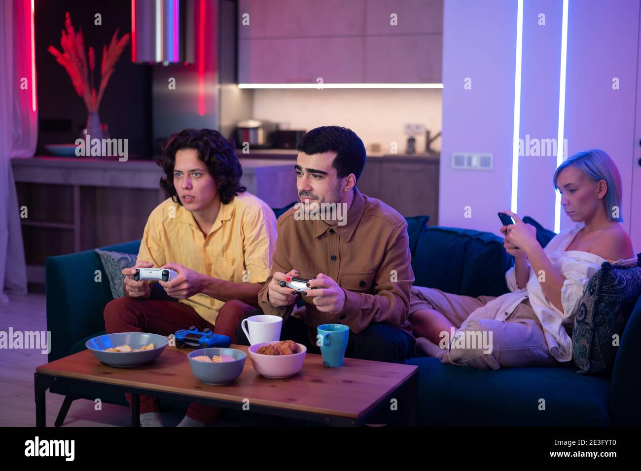 La compagnie des amis aiment se détendre sur un canapé jouant des jeux vidéo et s'amuser dans le moderne lumineux plat avec des couleurs de néon. Banque D'Images