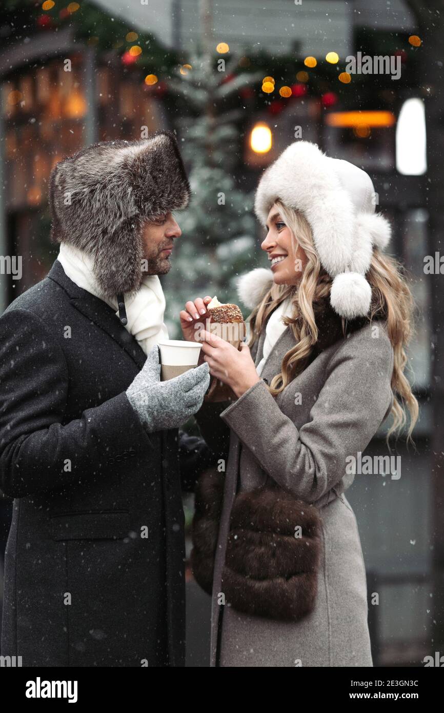 Un couple d'amateurs de mode se tenant sur la rue d'hiver par temps enneigé, souriant, buvant des boissons chaudes. Banque D'Images