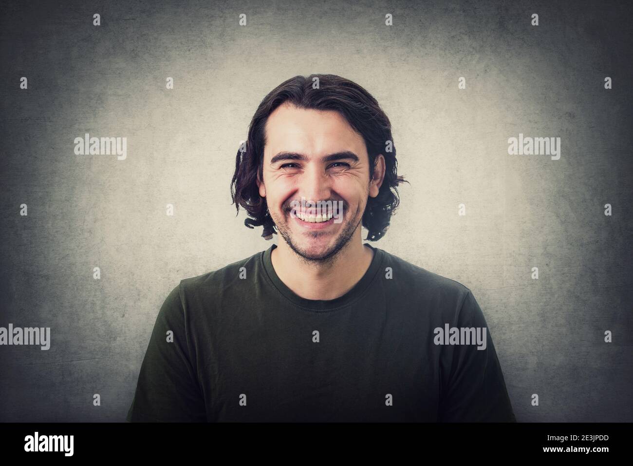 Portrait d'un jeune homme brunette souriant, cheveux longs et bouclés, aspect joyeux et riant à l'appareil photo isolé sur fond de mur gris. Gai et su Banque D'Images