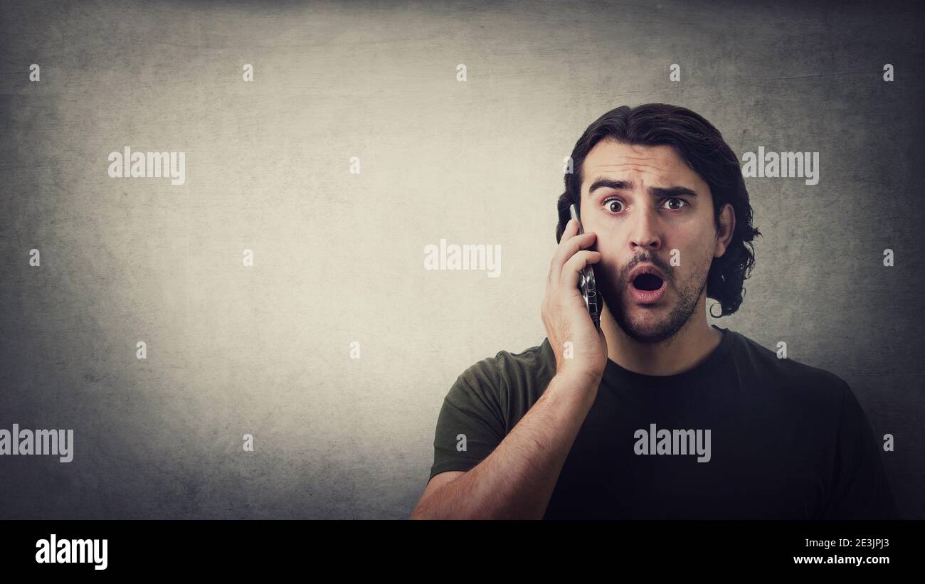 Un jeune homme choqué, au cours d'une conversation sur un téléphone portable, regardant avec de grands yeux et la bouche ouverte isolée sur fond de mur gris, avec un espace de copie. Stupéfait Banque D'Images