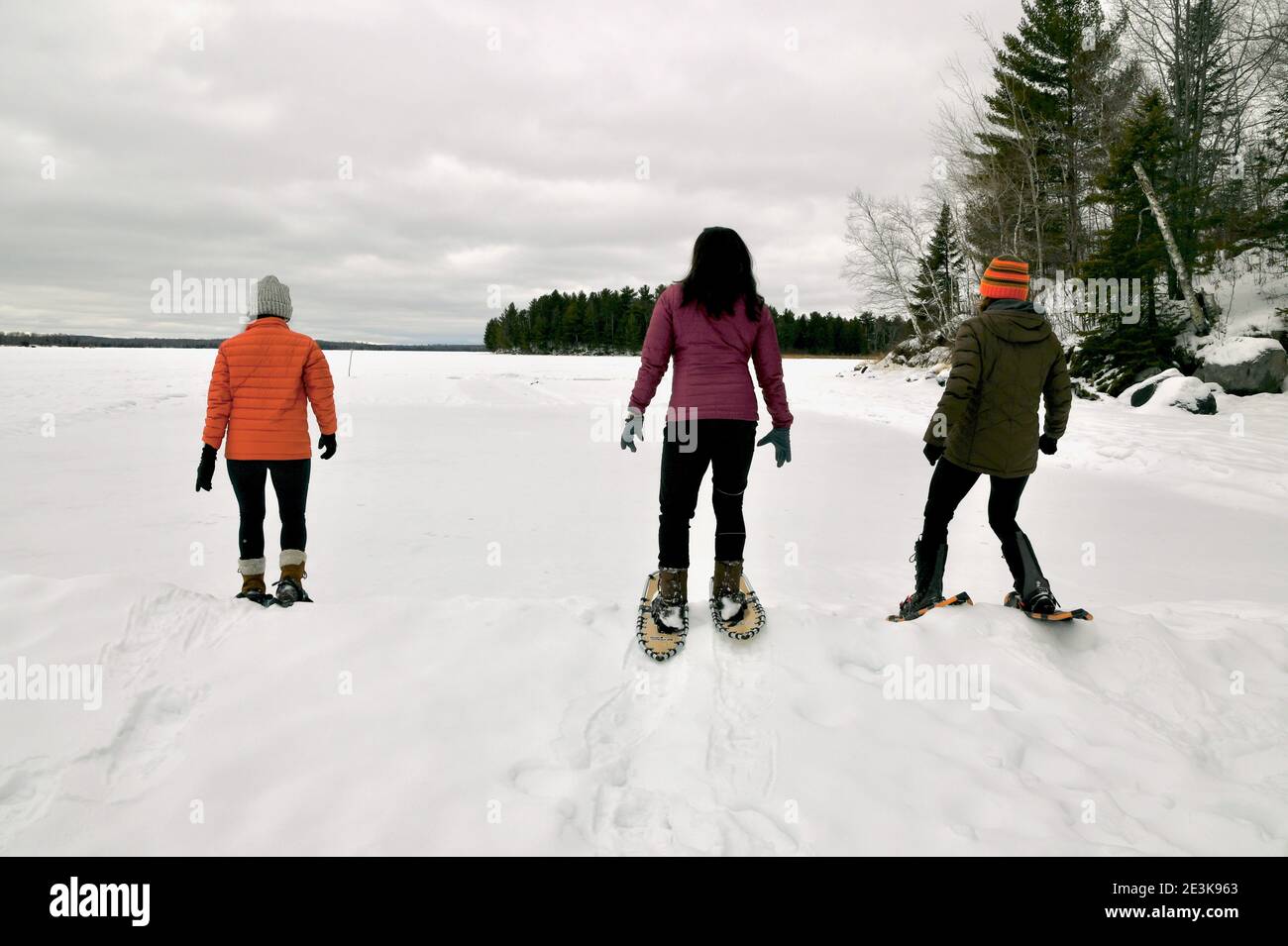 Les femmes raquette pendant l'hiver faisant des pistes de raquettes à travers le lac enneigé gelé avec un ciel hivernal nuageux et froid. Banque D'Images
