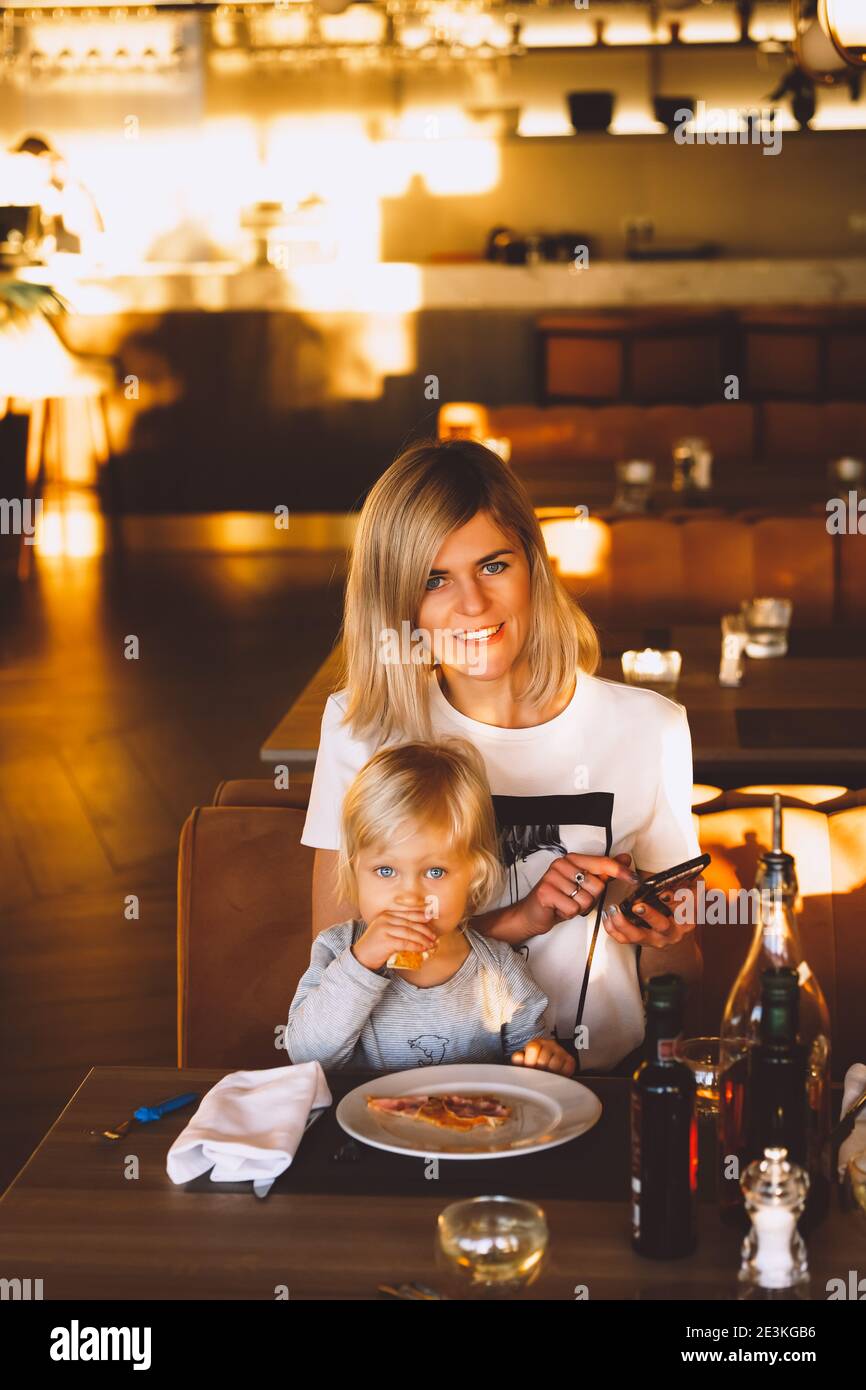 Jeune mère blonde se détendant dans un café intérieur ensoleillé messagerie texte avec son téléphone avec son petit enfant mignon manger de la pizza. Banque D'Images