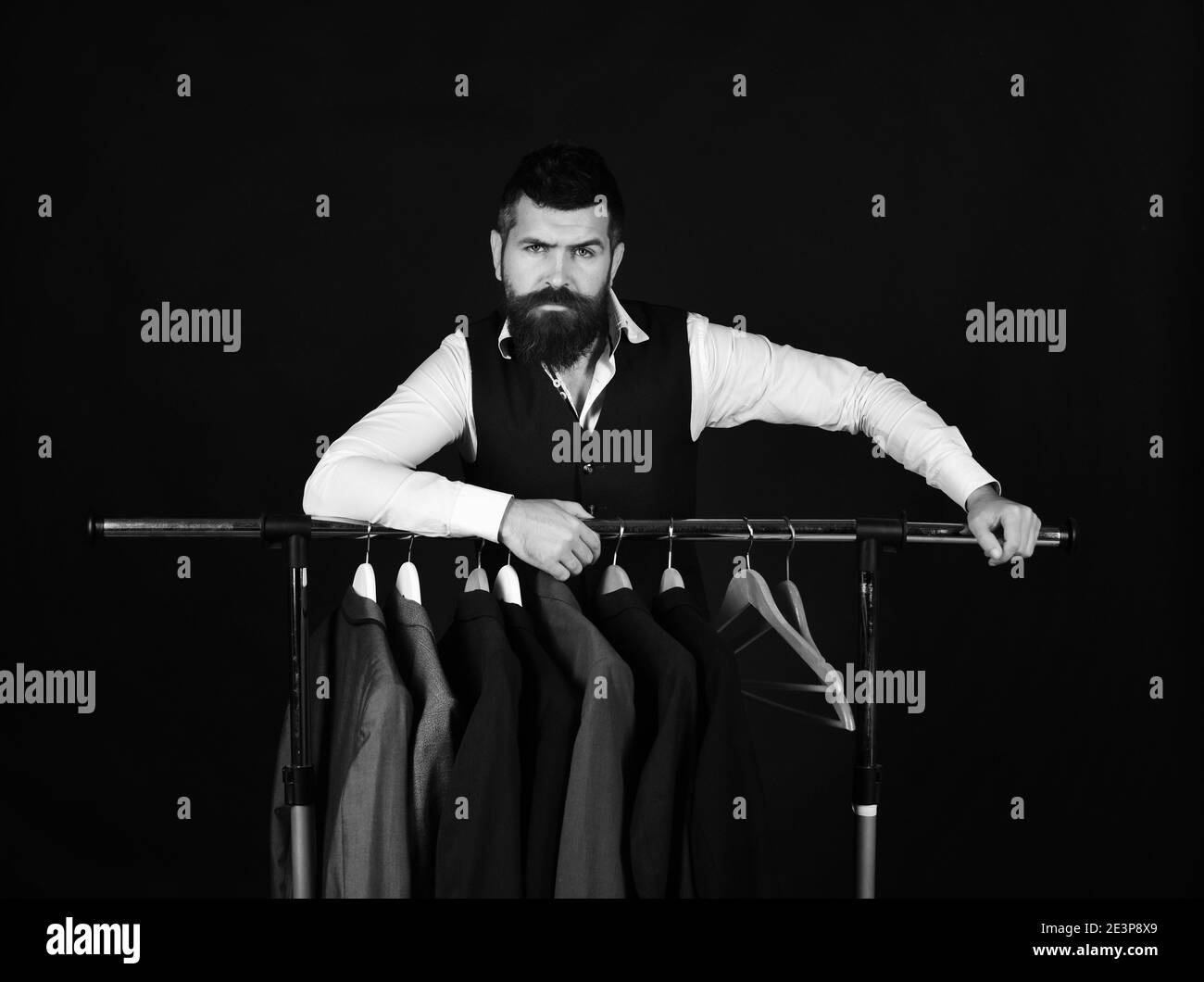 Mode et concept de style individuel. Homme avec barbe dans la veste près du porte-vêtements. Sur mesure avec un visage sérieux près des vestes personnalisées sur fond noir. Le designer s'appuie sur des cintres. Banque D'Images